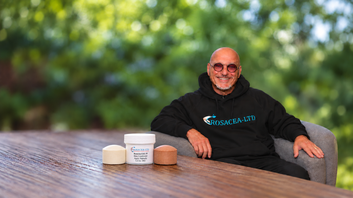 Howie Mandel smiling in a ROSACEA-LTD hoodie beside a ROSACEA-LTD jar and white and brown mineral discs on a wooden table.